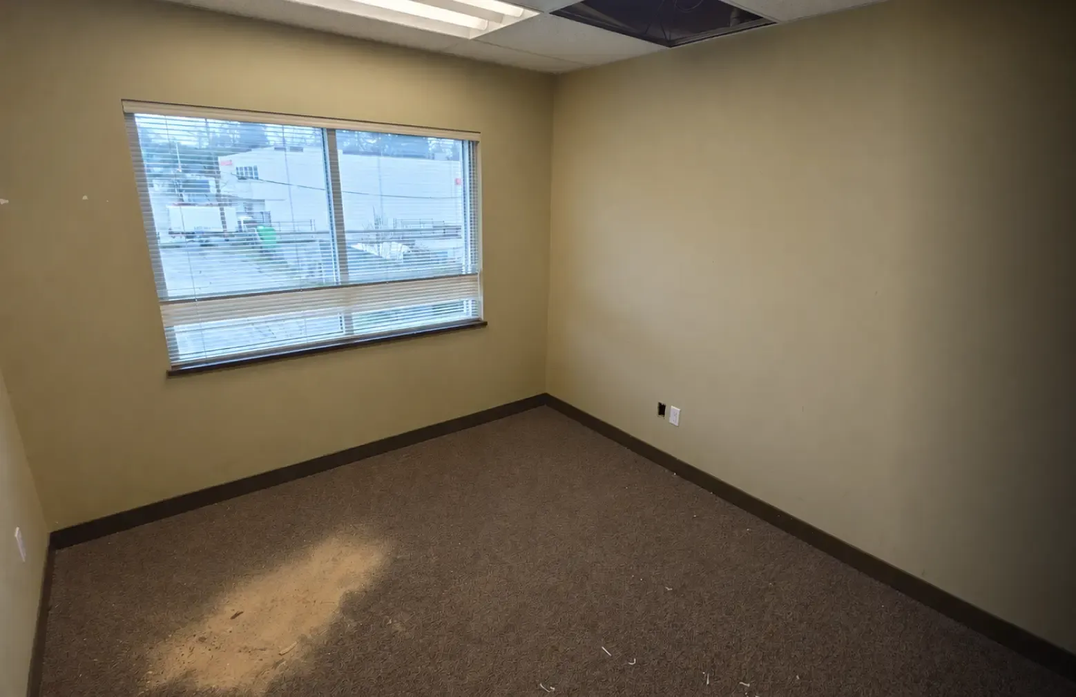 Before photo of a dated private office showing stained brown carpeting, beige walls, and missing ceiling tiles prior to a commercial tenant improvement by Pacific Coast Contracting.